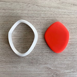 May include: A white plastic cookie cutter shaped like a rounded rectangle, next to a red fondant cutout of the same shape.