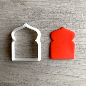 Puede incluir: Un cortador de galletas de plástico blanco con forma de puerta arqueada y un recorte de fondant rojo de la misma forma.