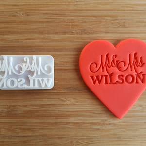 Könnte beinhalten: Ein weißer Plastik-Ausstecher mit dem Text "Mr & Mrs WILSON" und ein rotes herzförmiges Keks mit dem gleichen Text.