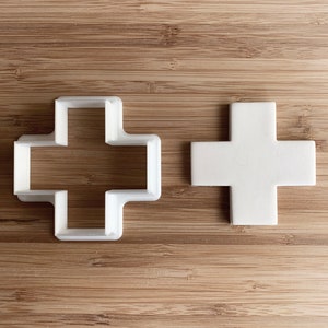 May include: Two white plastic cookie cutters in the shape of a plus sign. The cutter on the left is open and the cutter on the right is closed.