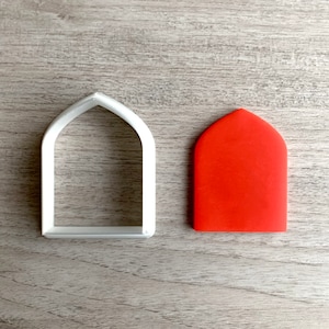 Puede incluir: Un cortador de galletas de plástico blanco con forma de ventana o puerta arqueada, junto a un trozo de material rojo liso, posiblemente fondant o masa. Los objetos están sobre una superficie de madera gris claro.