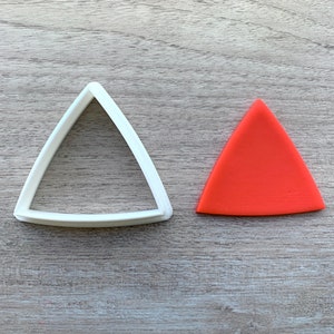 Puede incluir: Un cortador de galletas de plástico blanco en forma de triángulo y un recorte de galleta de triángulo rojo sobre una superficie de madera.