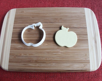 Peut inclure: Un emporte-pièce en forme de pomme en plastique blanc et une pomme en pâte d'amande jaune sur une planche à découper en bois.