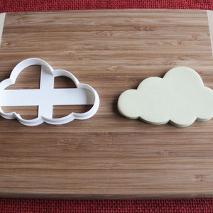 May include: A white plastic cloud cookie cutter and a light green fondant cloud on a wooden chopping board.