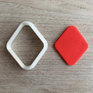 Op de afbeelding: Een witte plastic diamantvormige koekjesvormer en een rode diamantvormige koek op een houten achtergrond.