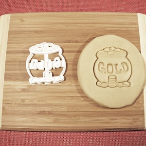 May include: A white plastic cookie cutter shaped like a pot of gold with the word "GOLD" on it. Next to it is a brown cookie dough cutout of the same shape.