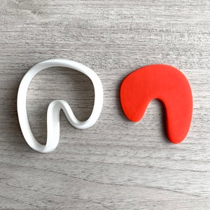May include: A white plastic cookie cutter in a curved shape, next to a solid orange cookie in the same shape. The cutter has a smooth, rounded design, and the cookie is a solid, vibrant orange color.