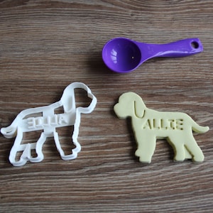 May include: Two white plastic dog-shaped cookie cutters, one with the name "Allie" and the other with the name "Ellie". A purple plastic measuring spoon is also in the image.