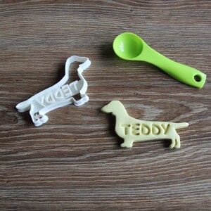 May include: Two white dachshund cookie cutters, one with the name "Teddy" on it, and a green plastic measuring spoon.