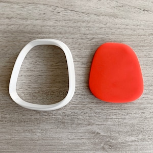 May include: A white plastic cookie cutter shaped like a rounded oval, next to a red oval-shaped piece of modeling clay.