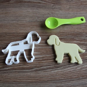 May include: Two dog-shaped cookie cutters, one white plastic and one yellow, on a brown wooden surface. A green plastic spoon is also in the image.