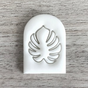Puede incluir: Molde de silicona blanco en forma de arco con un recorte de una hoja de monstera.