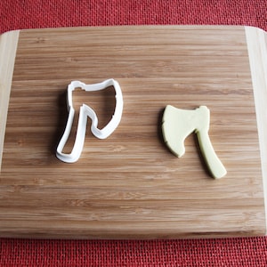 May include: Two axe-shaped cookie cutters on a wooden chopping board. One cutter is white plastic and the other is green.