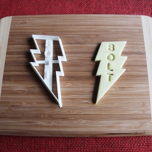 Può includere: Due stampini per biscotti a forma di fulmine. Uno è di plastica bianca e l'altro è giallo e ha la parola "BOLT" scritta su di esso.