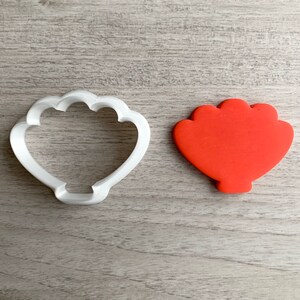 Puede incluir: Un cortador de galletas de plástico blanco con forma de concha, junto a una galleta roja con forma de concha.