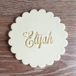 May include: A white, scalloped, round cookie cutter with the name "Elijah" embossed in a cursive font.