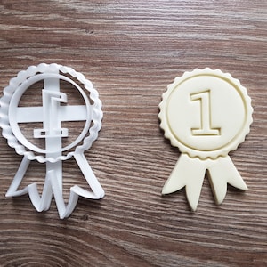 Op de afbeelding: Een witte plastic koekjesvormer en een gele koek in de vorm van een lint met het nummer "1" in het midden.