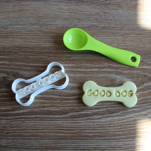 May include: A white plastic dog bone-shaped cookie cutter with the words "GOOD DOG" cut out. Next to it is a light yellow dog bone-shaped cookie with the words "GOOD DOG" imprinted. A green measuring spoon is also in the image.