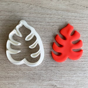 Puede incluir: Un cortador de galletas de plástico blanco con forma de hoja de monstera, junto a una hoja de monstera roja hecha de fondant.