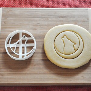 May include: A white plastic cookie cutter in the shape of a wolf howling at the moon, next to a plain round cookie with the same design pressed into it.