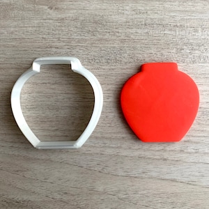 May include: A white plastic cookie cutter shaped like a vase, next to a red clay vase.