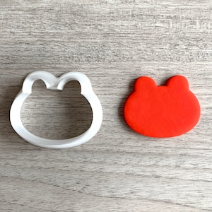 Può includere: Un tagliapasta di plastica bianca a forma di rana, accanto a un biscotto rosso a forma di rana.