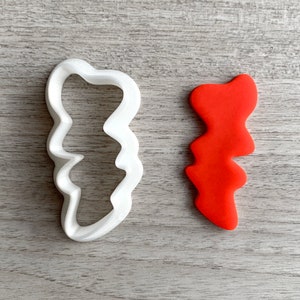 May include: A white plastic cookie cutter shaped like a jagged leaf, next to a red leaf-shaped piece of modeling clay.
