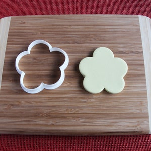 May include: A white plastic flower-shaped cookie cutter and a pale yellow fondant flower on a wooden chopping board.