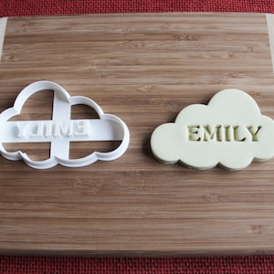 Può includere: Due stampini per biscotti in plastica bianca a forma di nuvola, uno con il nome "Emily" tagliato e l'altro con il nome "Jimmy" tagliato. Lo stampino "Emily" è su un tagliere di legno ed è riempito con un impasto verde chiaro.