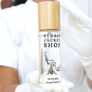 May include: A clear cylindrical glass spray bottle with a light-coloured wooden cap. The label features the text STRAIT JACKET SHOP, a graphic illustration of a medical chair, and the words MEMORIES and Dental Office printed in black ink.