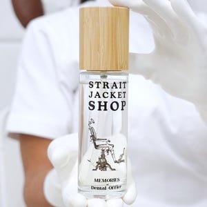 May include: A clear glass spray bottle with a light wood cap. The label features the black text STRAIT JACKET SHOP, a graphic illustration of a vintage dental chair, and the text MEMORIES Dental Office printed in black on the front surface.