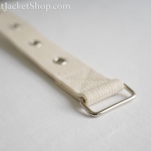 Può includere: Primo piano di un cinturino in tela bianca con una fibbia in metallo argentato. Il cinturino ha diversi occhielli argentati disposti uniformemente.