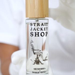 May include: A clear glass spray bottle with a light brown bamboo cap. The label features the black text "STRAIT JACKET SHOP" above a detailed illustration of a vintage dental chair, with the text "MEMORIES of Dental Office" printed below.