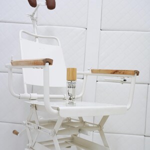 May include: A white metal chair with wooden armrests and a headrest featuring two brown pads. A small clear glass bottle with a bamboo cap sits on the seat, labelled with the text Strait Jacket Shop and an illustration of a chair.