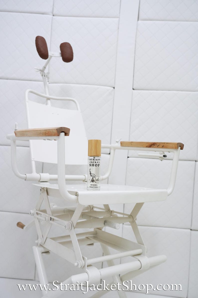 May include: A white metal chair with wooden armrests and a headrest featuring two brown pads. A small clear glass bottle with a bamboo cap sits on the seat, labelled with the text Strait Jacket Shop and an illustration of a chair.