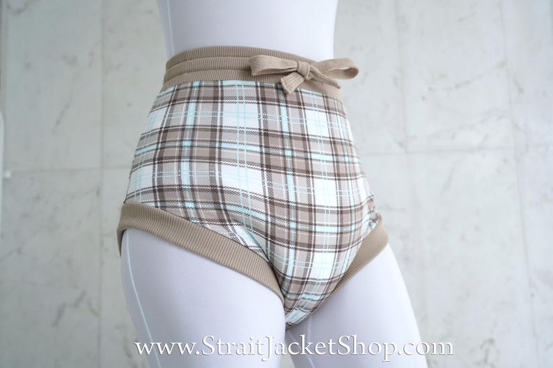May include: High-waisted shorts with a plaid pattern in shades of brown, blue, and white. The waistband and leg openings are trimmed with a solid tan fabric. A small bow detail is centered on the front of the waistband. The shorts are displayed on a mannequin.