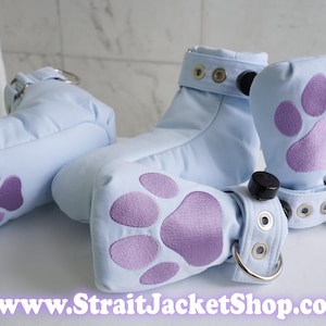 May include: Light blue padded boots featuring purple paw print appliques on the front and sides. Each boot includes a strap with metal eyelets, a silver-toned D-ring, and a black circular fastener. The text www.StraitJacketShop.com is visible.