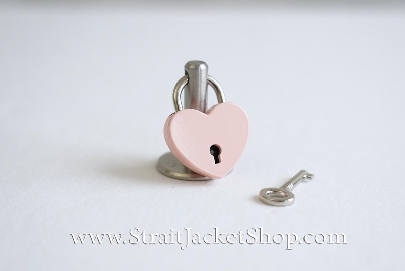 May include: A small pink heart-shaped padlock with a silver key on a white background. The padlock is attached to a silver stand.