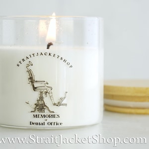 May include: A white candle in a clear glass jar with a lit wick. The label features a black line drawing of a vintage dental chair, the text STRAITJACKETSHOP, and the words MEMORIES of Dental Office. A light wood lid sits beside the jar.