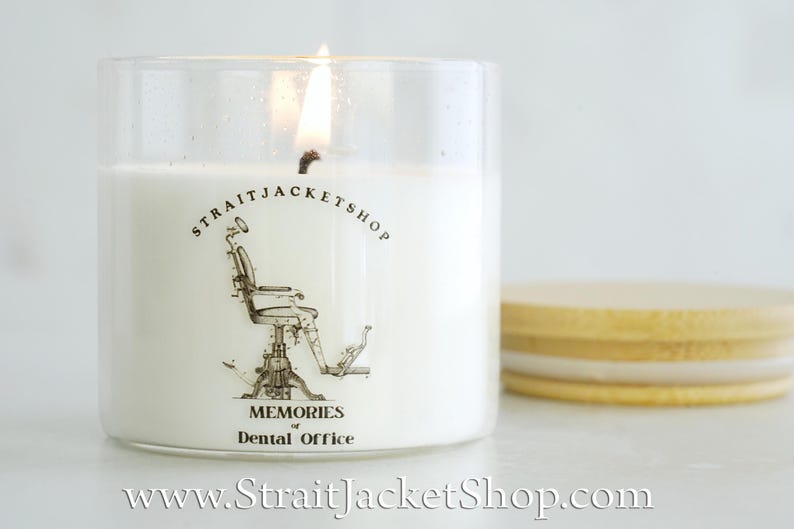 May include: A white candle in a clear glass jar with a lit wick. The label features a black line drawing of a vintage dental chair, the text STRAITJACKETSHOP, and the words MEMORIES of Dental Office. A light wood lid sits beside the jar.