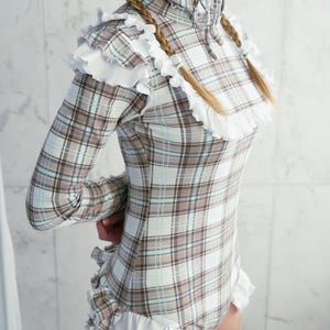 May include: A long-sleeved plaid bodysuit in shades of brown, white, and light blue, trimmed with white ruffles. The garment has a high collar and a zipper closure. The website address www.StraitJacketShop.com is visible at the bottom.