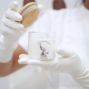 May include: A white wax candle in a clear glass jar with a bamboo lid. The label features a black and white illustration of a dental chair with the text STRAITJACKETSHOP, MEMORIES, and Dental Office printed in black ink on the front of the jar.
