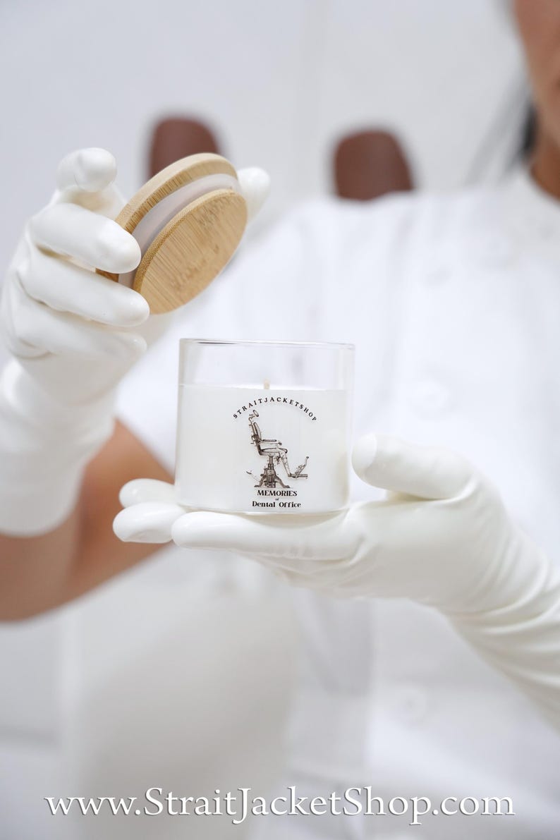 May include: A white wax candle in a clear glass jar with a bamboo lid. The label features a black and white illustration of a dental chair with the text STRAITJACKETSHOP, MEMORIES, and Dental Office printed in black ink on the front of the jar.