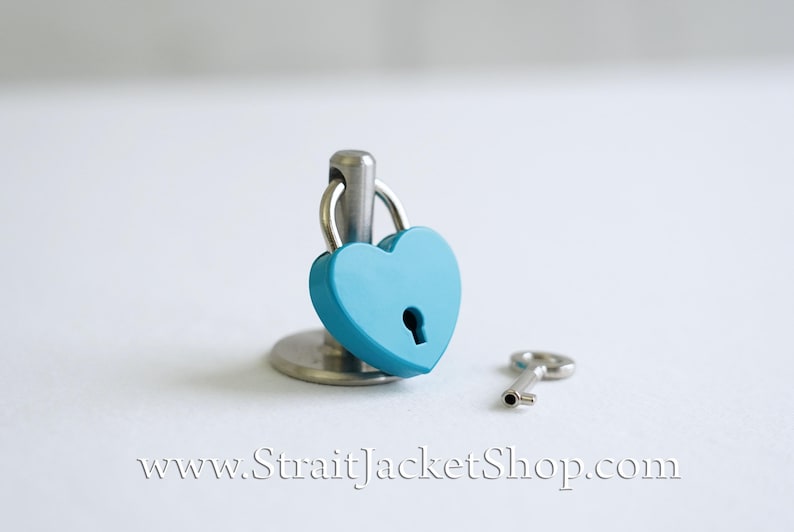 May include: A small, heart-shaped padlock with a key. The padlock is blue and the key is silver. The padlock is on a small stand.