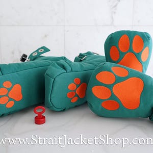 May include: A set of teal fabric mittens featuring bright orange paw print appliques on the palms. Each mitten includes a wrist strap with silver metal grommets, a black adjustment knob, and a small red plastic key accessory is placed nearby.