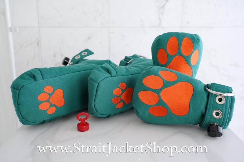 May include: A set of teal fabric mittens featuring bright orange paw print appliques on the palms. Each mitten includes a wrist strap with silver metal grommets, a black adjustment knob, and a small red plastic key accessory is placed nearby.