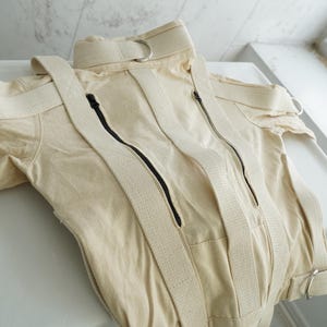 Peut inclure: Vêtement beige avec des fermetures éclair noires et des sangles crème. Le vêtement a un col montant et des détails d'anneaux métalliques. Le tissu semble être un mélange de coton ou de lin robuste. Le design est utilitaire et structuré.