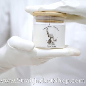 May include: A clear glass candle jar with a light wood lid, filled with white wax. The label features a black line drawing of a dental chair, the text STRAITJACKETSHOP, and the words MEMORIES of Dental Office printed in black ink.