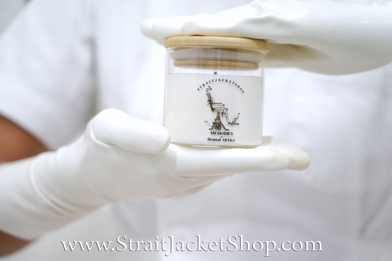 May include: A clear glass candle jar with a light wood lid, filled with white wax. The label features a black line drawing of a dental chair, the text STRAITJACKETSHOP, and the words MEMORIES of Dental Office printed in black ink.