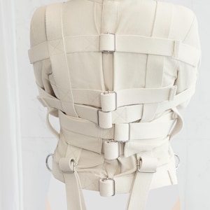 May include: A white canvas straitjacket with multiple straps and buckles. The straps are adjustable and have metal buckles. The straitjacket is designed to restrain the arms and torso. The text "www.StraitjacketShop.com" is visible at the bottom of the image.